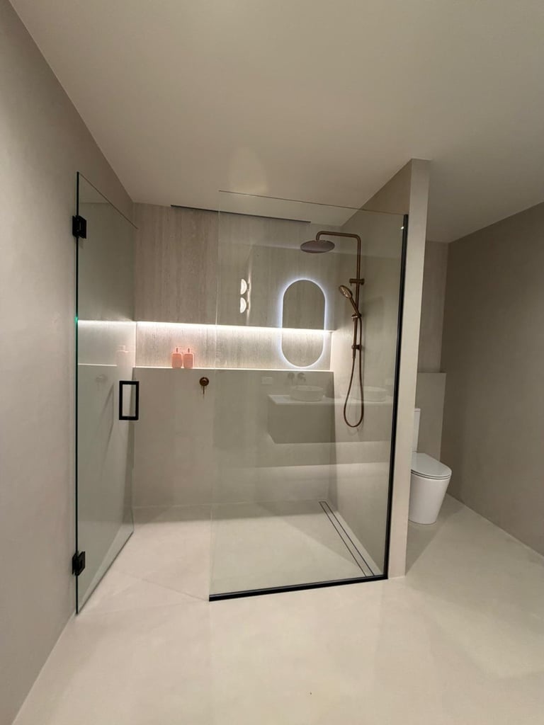Modern bathroom shower with frameless glass enclosure, rainfall showerhead, and illuminated oval mirror in neutral tones