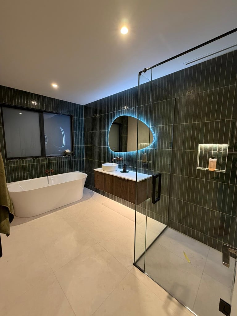 Modern bathroom with white freestanding bathtub, dark green tiled walls, cloud-shaped mirror with blue lighting, and glass shower enclosure