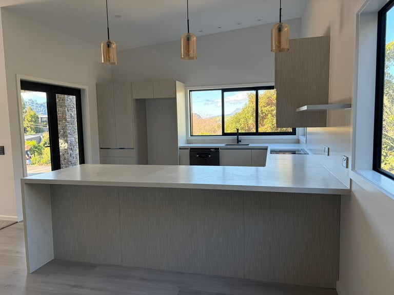 Modern kitchen with gray cabinetry, white countertops, island, brass pendant lights, and large windows overlooking autumn landscape.